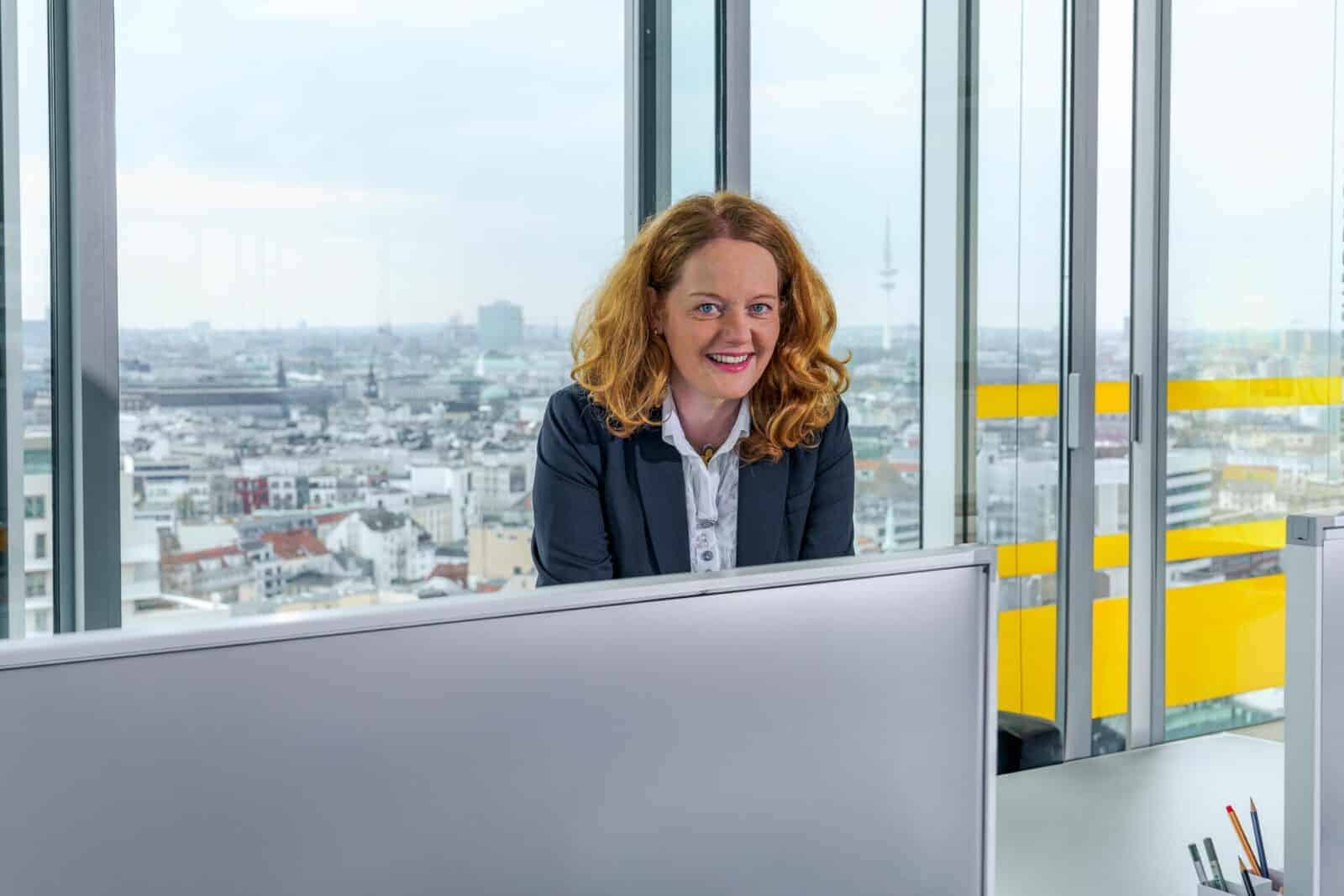 Businessfotografie Hamburg – sympathische Frau in hellem Büro mit Blick auf die Hamburger Skyline durch Glasfassade