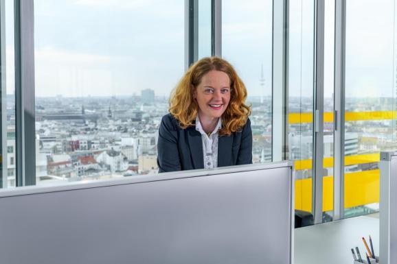 Businessfotografie Hamburg – sympathische Frau in hellem Büro mit Blick auf die Hamburger Skyline durch Glasfassade