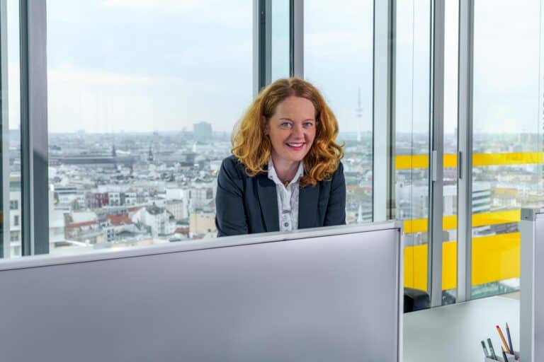 Businessfotografie Hamburg – Frau im Büro mit Skyline Businessfotografie Hamburg – sympathische Frau in hellem Büro mit Blick auf die Hamburger Skyline durch Glasfassade