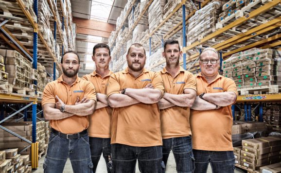 industriefoto-mitarbeiterportrait-lagerarbeiter Industriefotografie Hamburg – Team aus Lagerarbeitern in orange Poloshirts im modernen Logistiklager