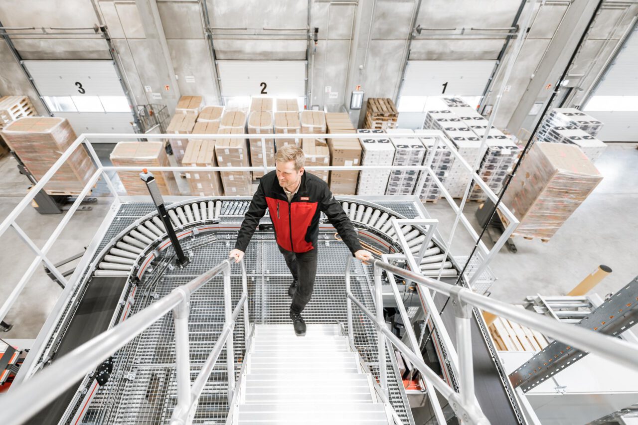 Intralogistik Fotografie – moderner Treppenaufgang in Lagerhalle mit hohen Regalen, Architektur und Struktur industrieller Logistikräume