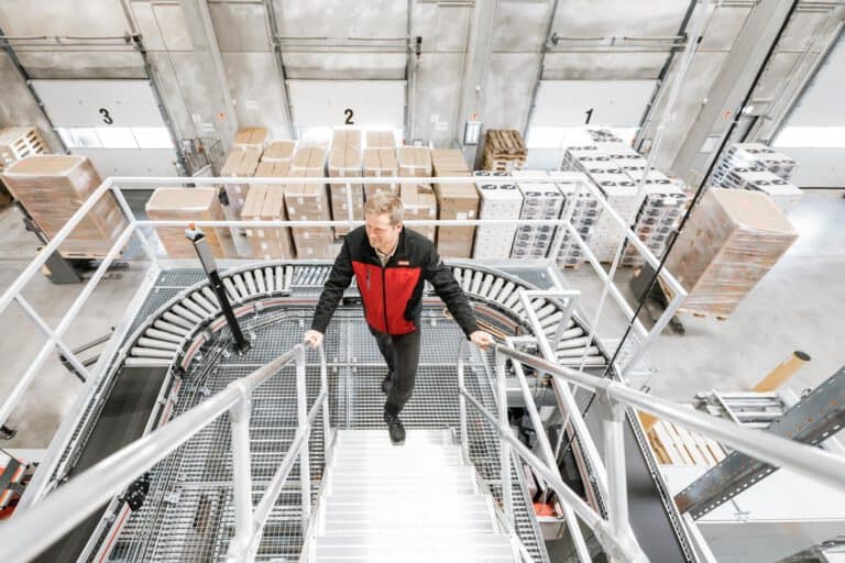 Lagerhalle Treppenaufgang – Intralogistik Fotografie Hamburg Intralogistik Fotografie – moderner Treppenaufgang in Lagerhalle mit hohen Regalen, Architektur und Struktur industrieller Logistikräume