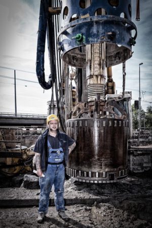Portrait Baustellenarbeiter – Industriefotografie Baustellenarbeiter vor schwerem Bohrgerät – authentisches Industriefotografie-Portrait