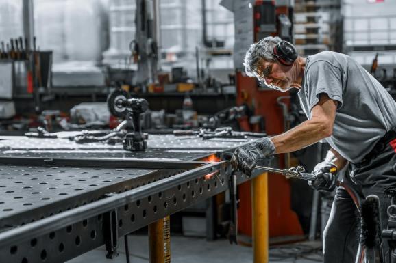 Metallbau Fotografie – Mitarbeiter in industrieller Produktion, Bearbeitung von Metallteilen an moderner Maschine mit Funkenflug